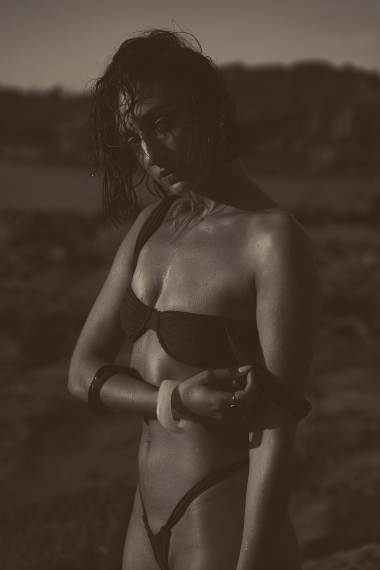 Woman in a bikini stands in sepia tones against an outdoor background, accessorized with bangle bracelets and rings.