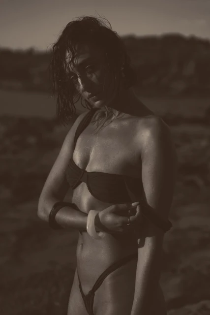 Woman in a bikini stands in sepia tones against an outdoor background, accessorized with bangle bracelets and rings.
