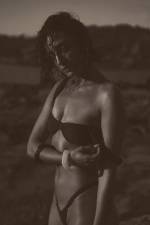 Woman in a bikini stands in sepia tones against an outdoor background, accessorized with bangle bracelets and rings.