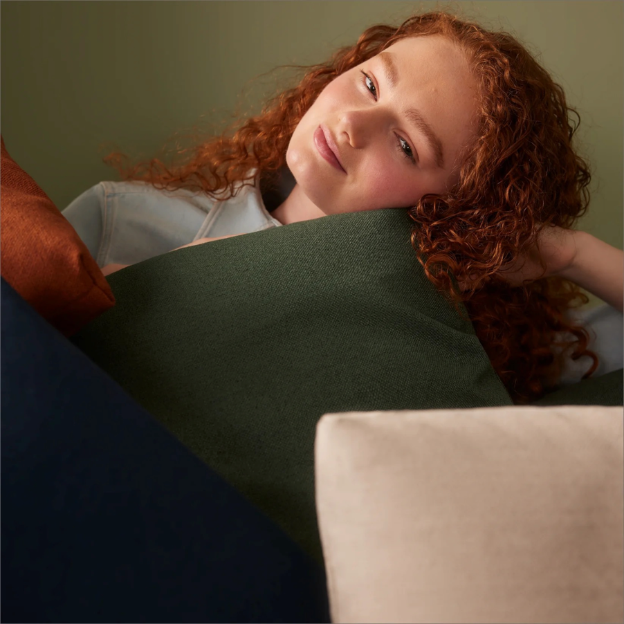 Red-haired woman in a light blue top reclines with head resting on a green pillow, surrounded by decorative pillows against a green wall.