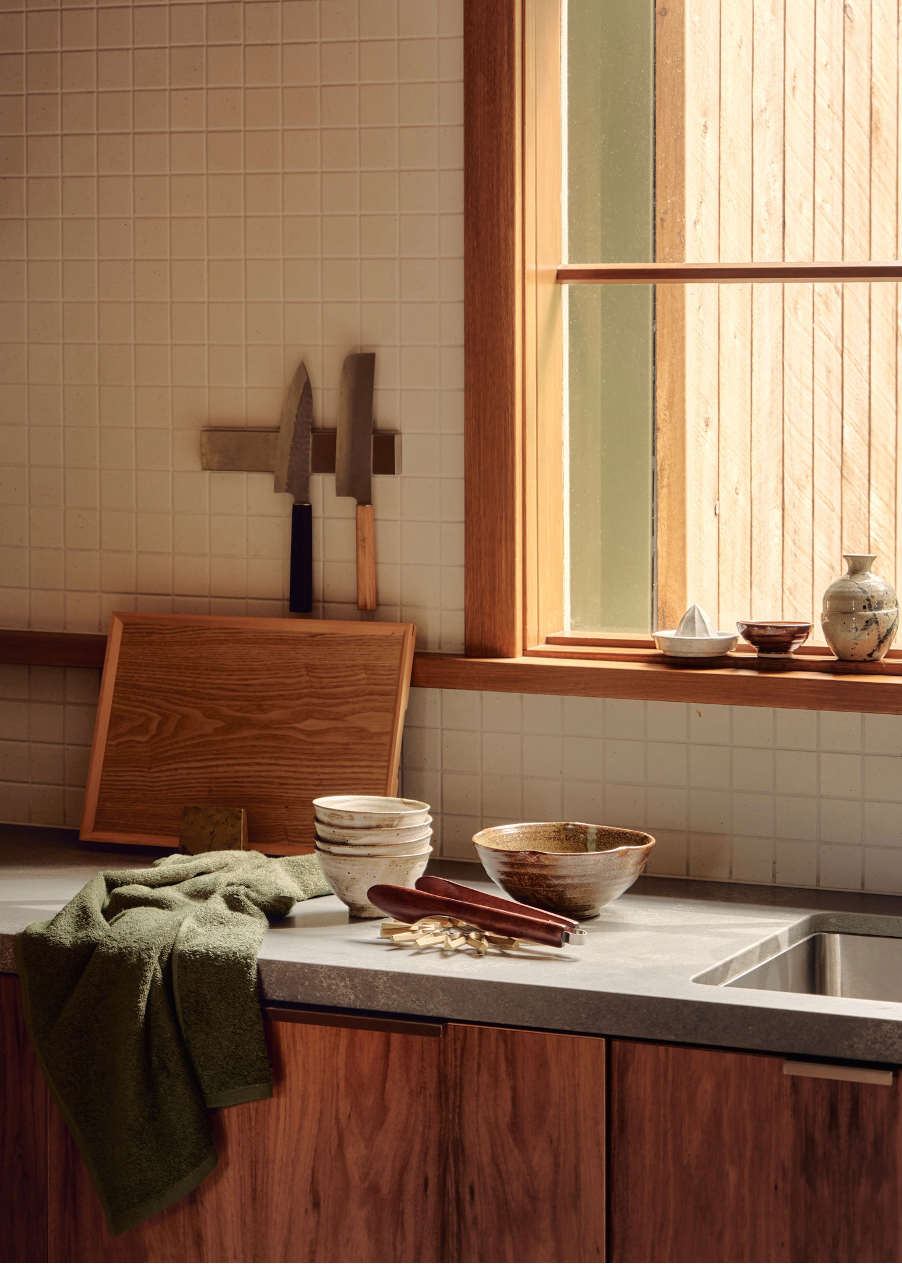 Kitchen still life with ceramic bowls, wooden utensils, knives, and a green towel on a gray countertop with a wood cabinet base.