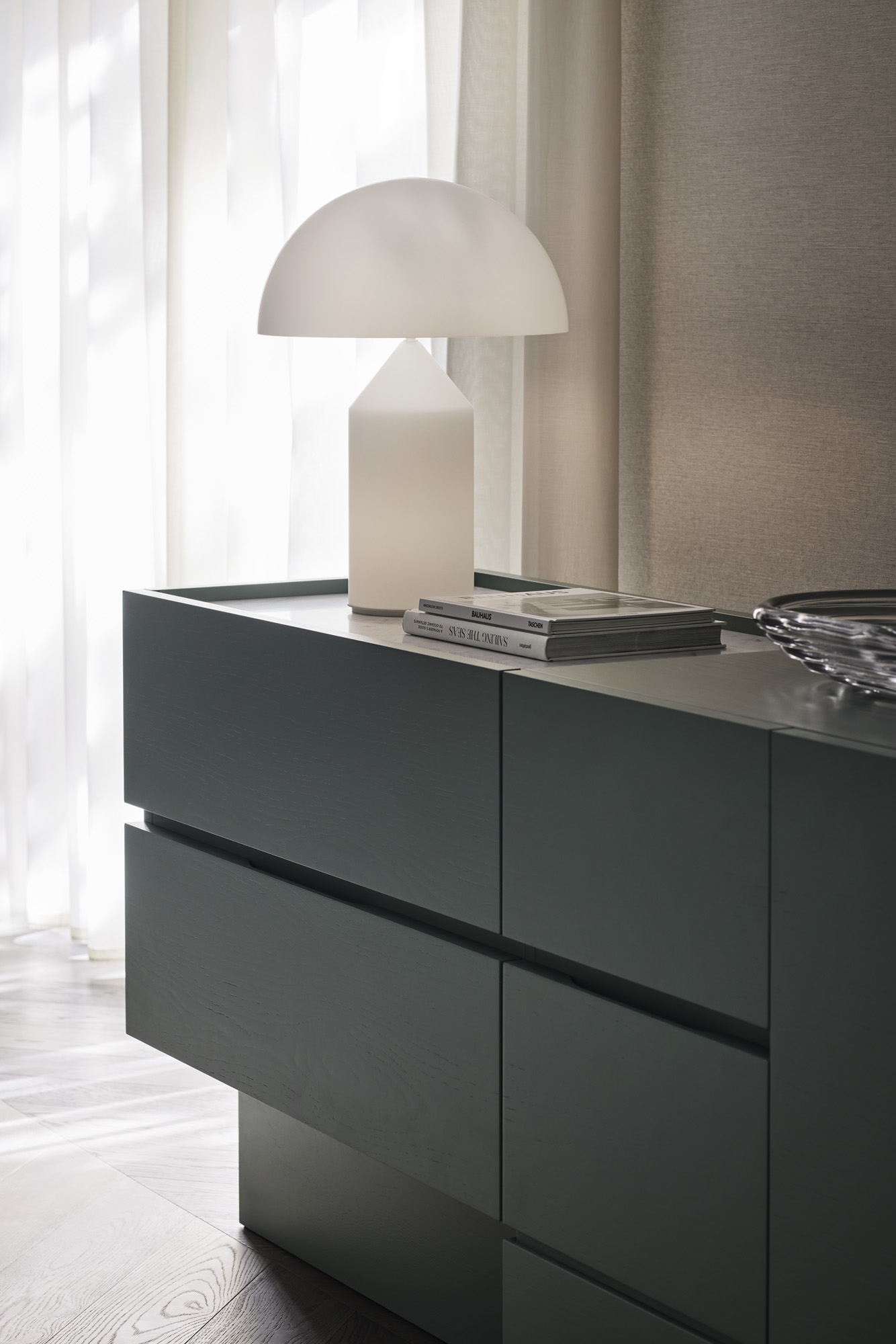 Interior scene features a modern grey commode with drawers, complemented by a white lamp and books, all set against sheer curtains.