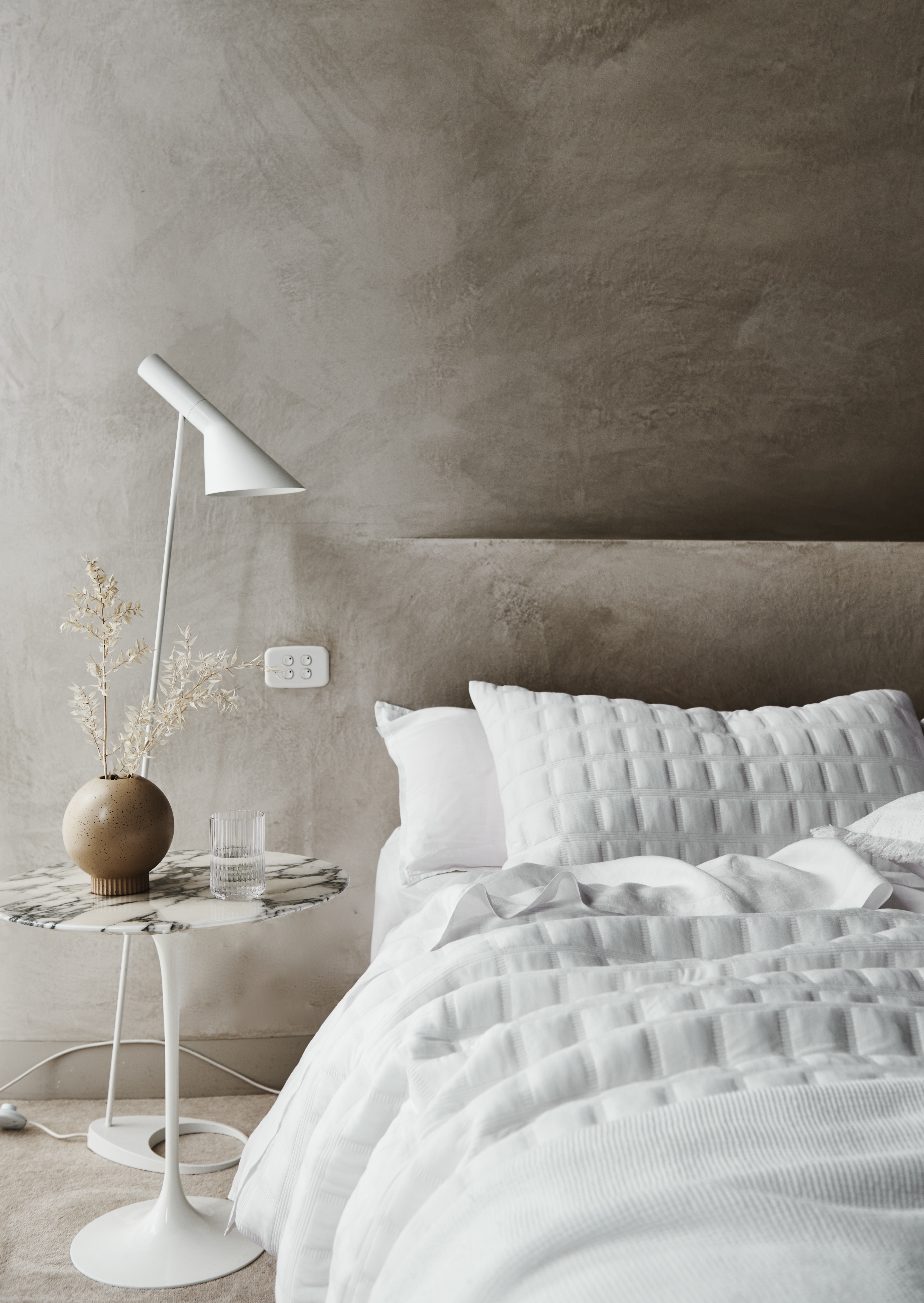 Bedroom interior featuring a white bed with textured bedding, marble side table with a lamp, vase and glass, against a textured grey wall.