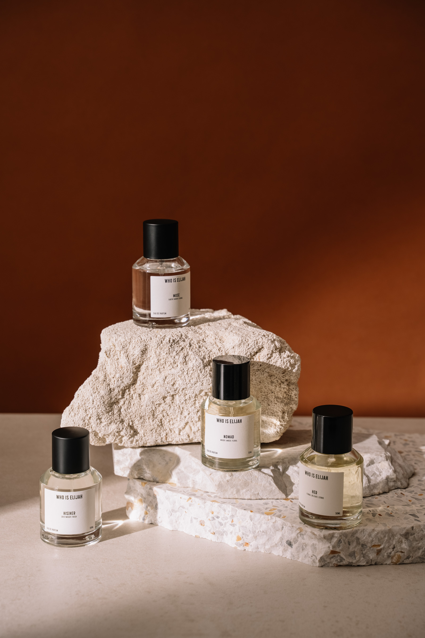 Four glass perfume bottles with black caps are arranged on white stone blocks against a warm brown background, creating a minimalist display.
