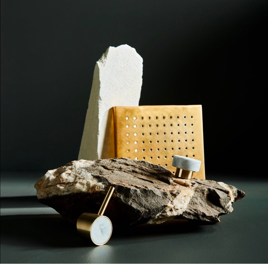 Still life of rocks, a gold colored card, and cosmetic container on a dark gray surface, with a dark gray background.