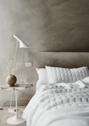Minimalist bedroom interior with white quilted bedding against a textured taupe plaster wall and a marble side table with a white lamp.