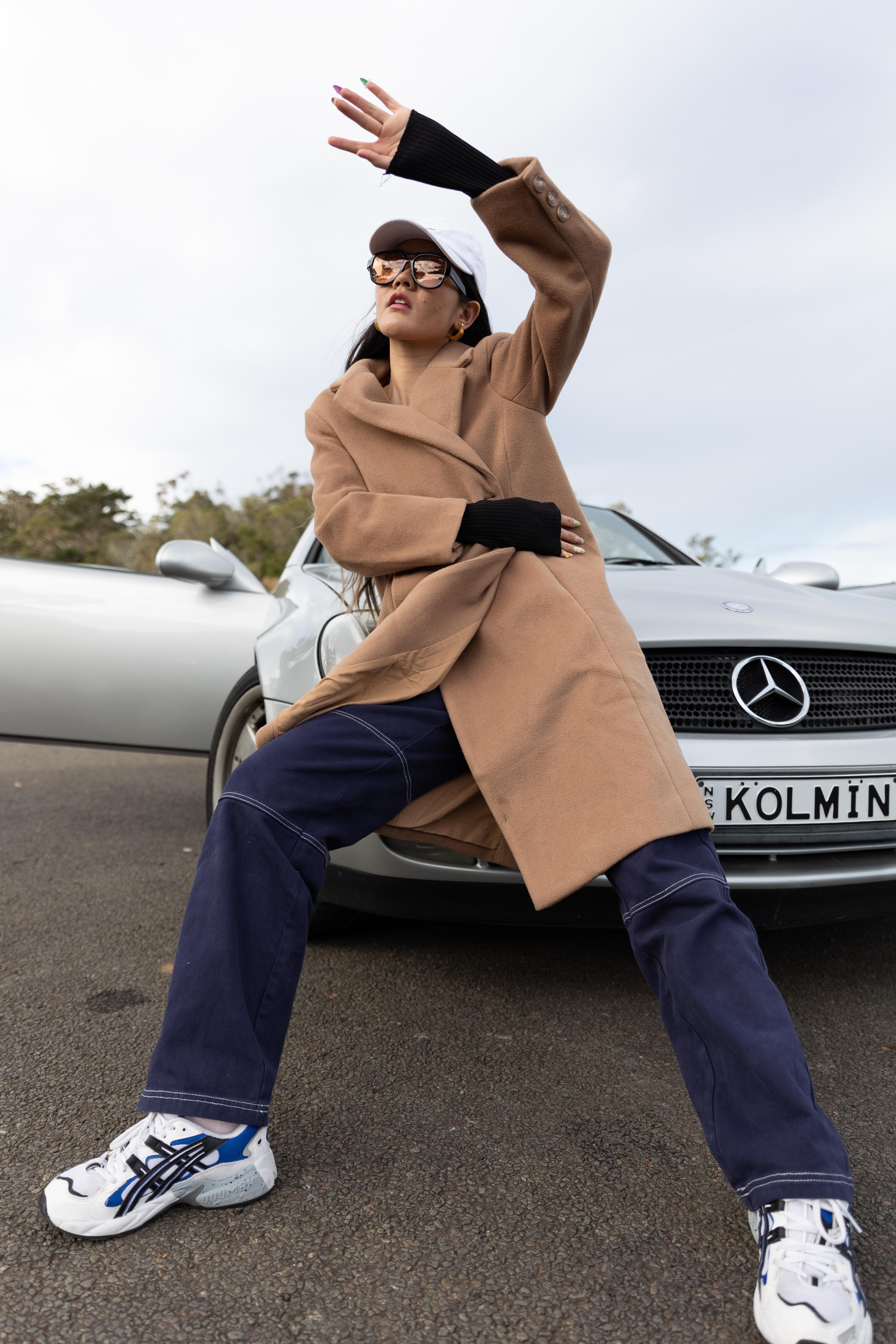 A woman poses by a silver car on an overcast day, wearing a beige coat, jeans, sneakers, sunglasses and a white cap.