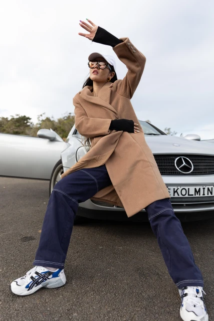 A woman poses by a silver car on an overcast day, wearing a beige coat, jeans, sneakers, sunglasses and a white cap.