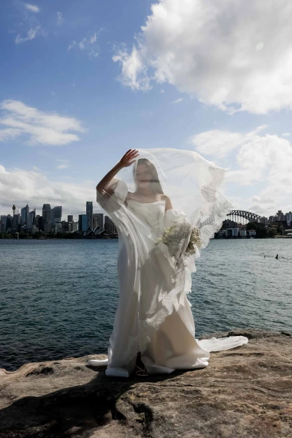 A woman in a wedding dress and veil stands on a rocky shore with a bouquet, with a cityscape and ocean in the background on a sunny day.