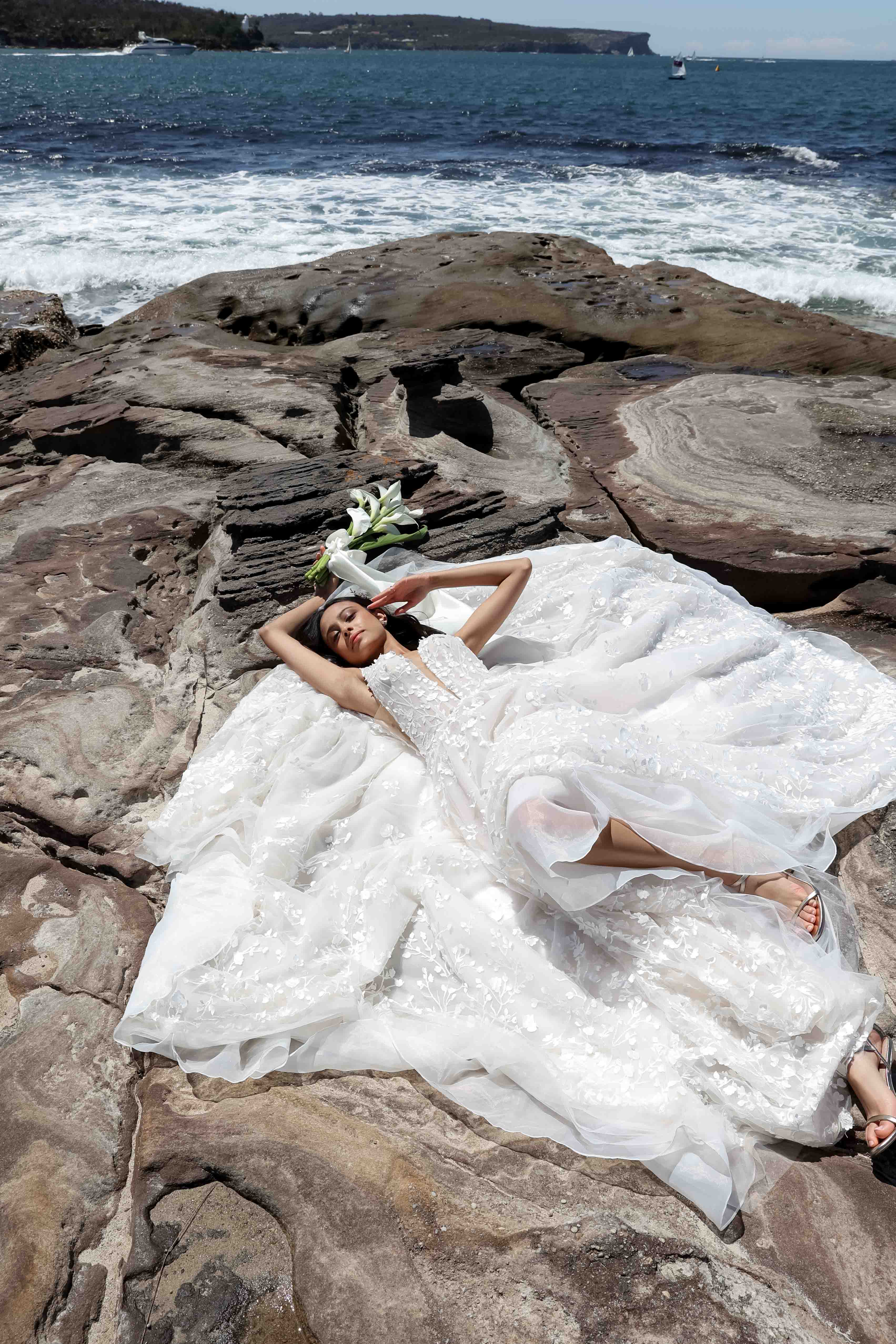 A woman in a wedding dress poses on a rocky shore next to white flowers with a seascape background under bright sunlight. 