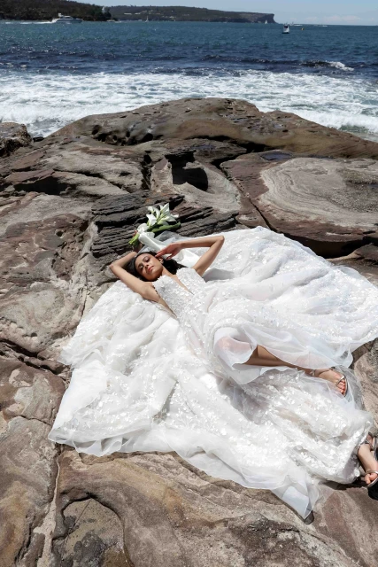 A woman in a wedding dress poses on a rocky shore next to white flowers with a seascape background under bright sunlight. 