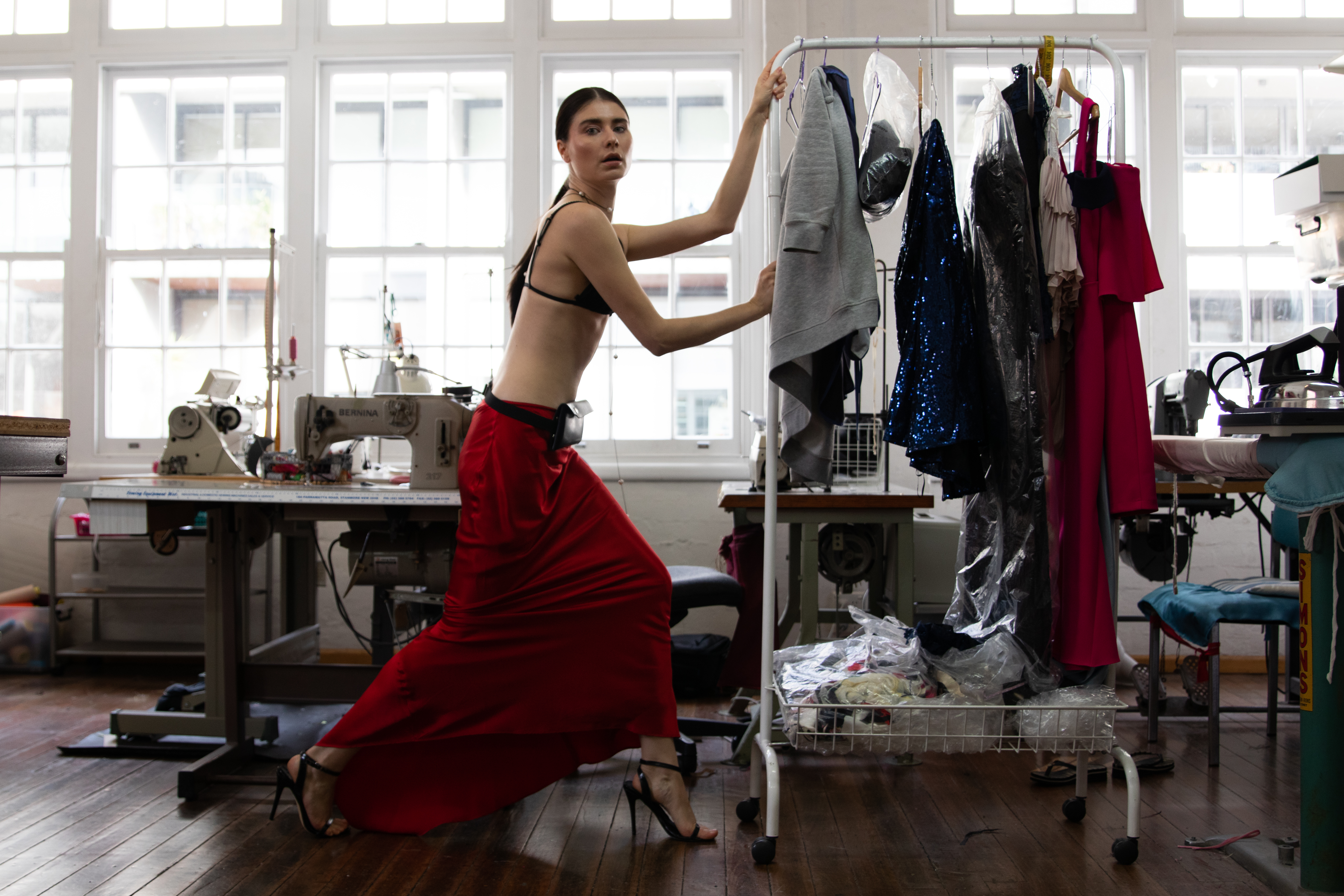 Brunette model poses in a red skirt and bra top in a sewing studio, surrounded by sewing machines, clothes racks, and windows.