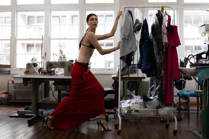 Brunette model poses in a red skirt and bra top in a sewing studio, surrounded by sewing machines, clothes racks, and windows.