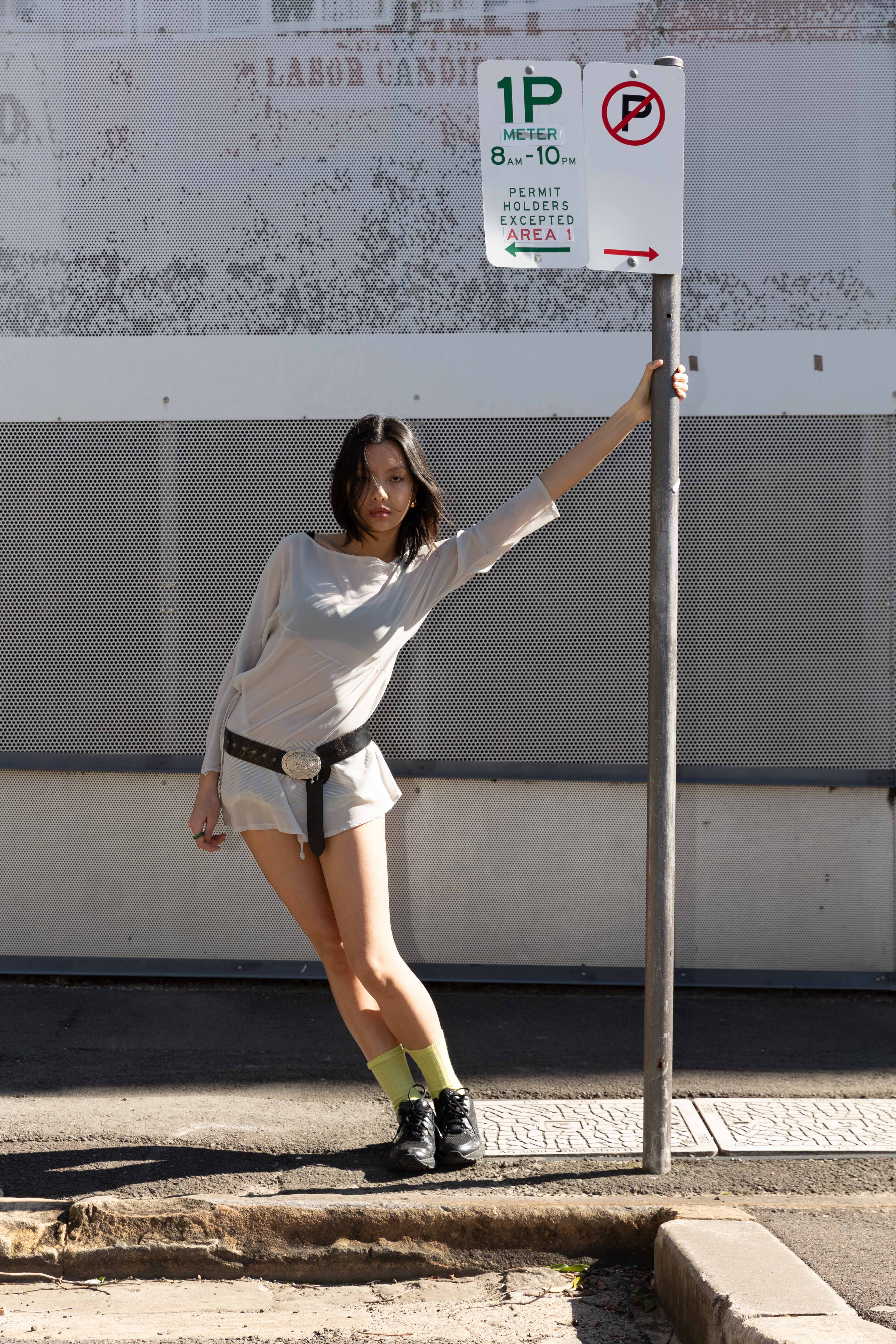 A slender woman posing in front of a weathered wall with a parking sign, showcasing a casual yet stylish fashion ensemble.