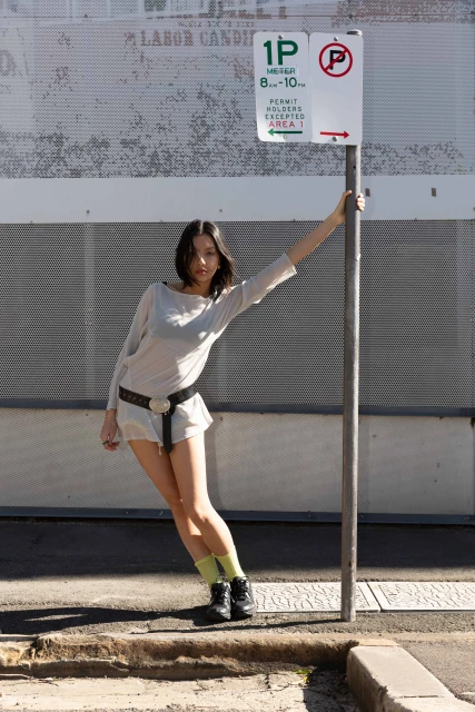 A slender woman posing in front of a weathered wall with a parking sign, showcasing a casual yet stylish fashion ensemble.
