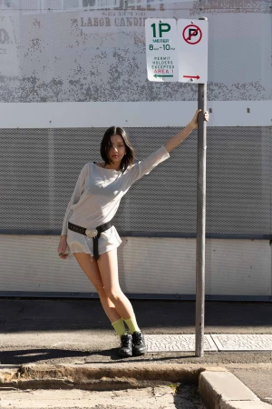 A slender woman posing in front of a weathered wall with a parking sign, showcasing a casual yet stylish fashion ensemble.