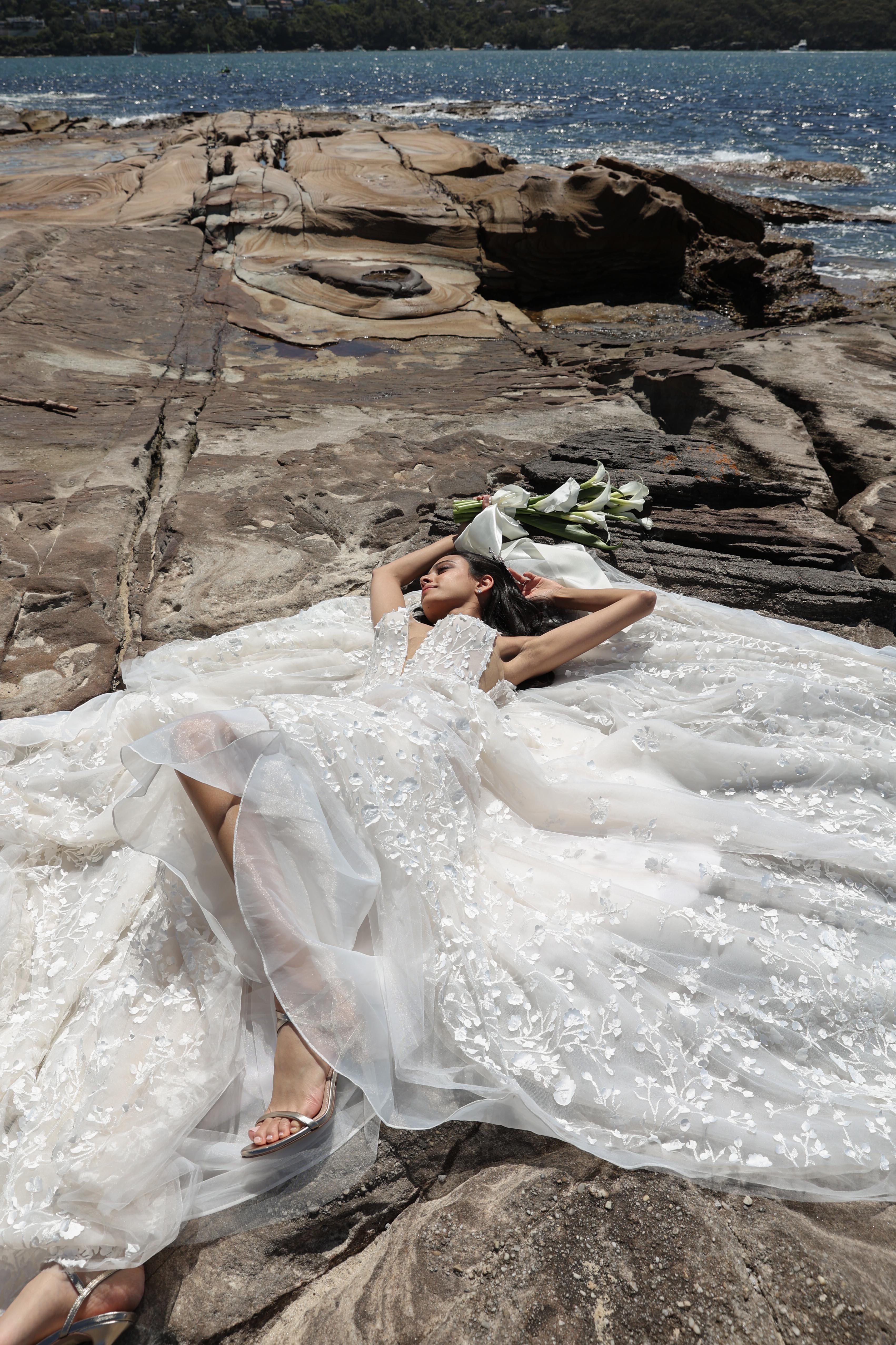 An adult woman in a white gown poses on rocky beach with white flowers, with a background of ocean and trees on the coast. 