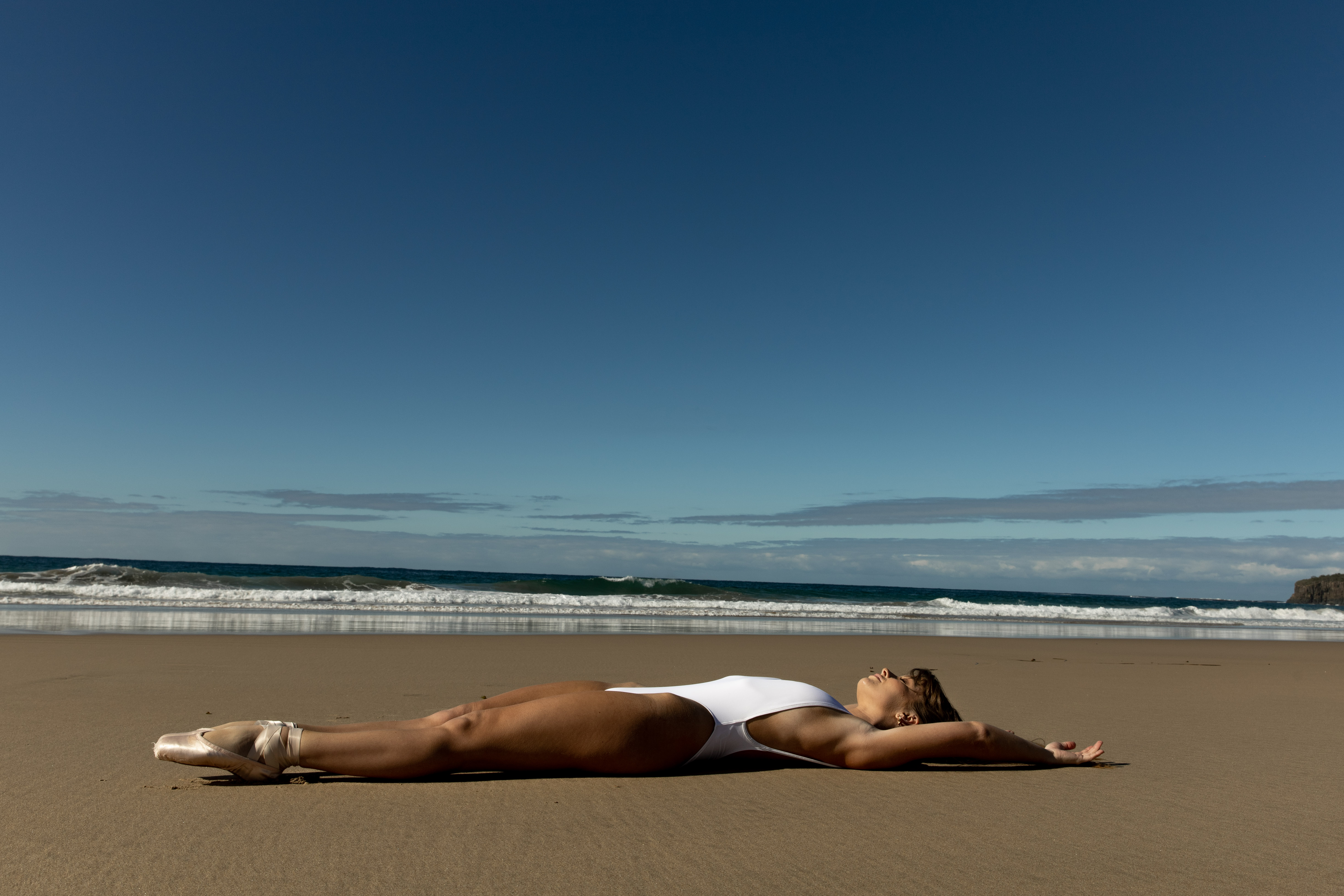 Woman in a white swimsuit and ballet shoes lies on a sandy beach with a blue sky and ocean in the background during daylight.
