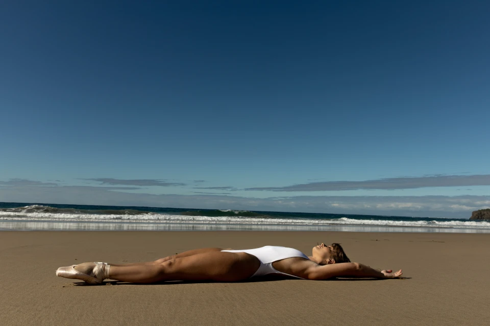 Woman in a white swimsuit and ballet shoes lies on a sandy beach with a blue sky and ocean in the background during daylight.