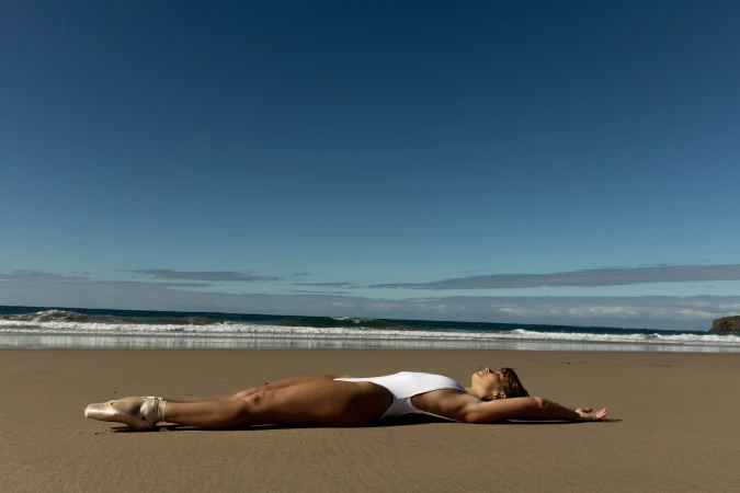 Woman in a white swimsuit and ballet shoes lies on a sandy beach with a blue sky and ocean in the background during daylight.