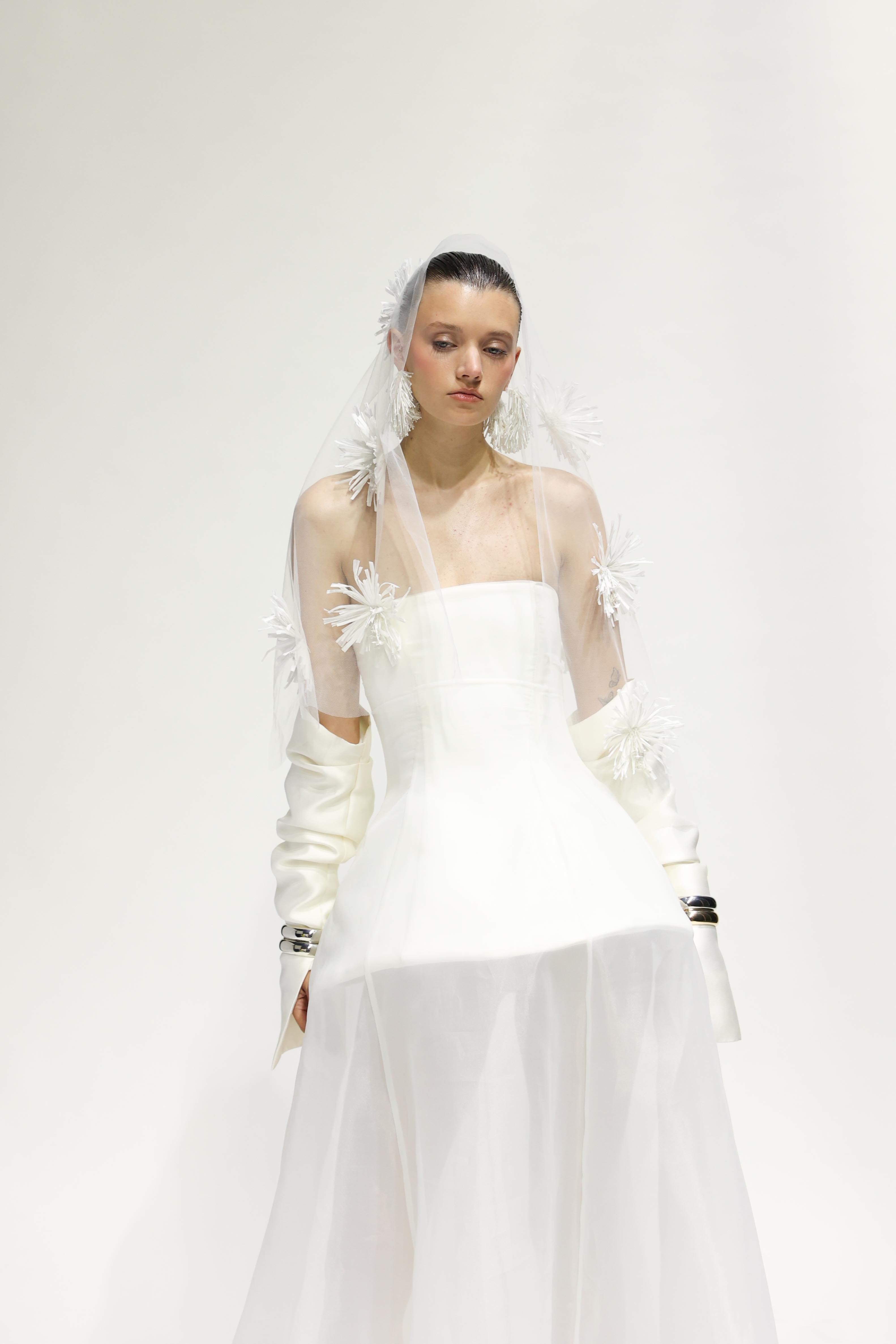 A woman poses in an off-shoulder white wedding dress and tulle veil with white flower accents against a plain white backdrop.