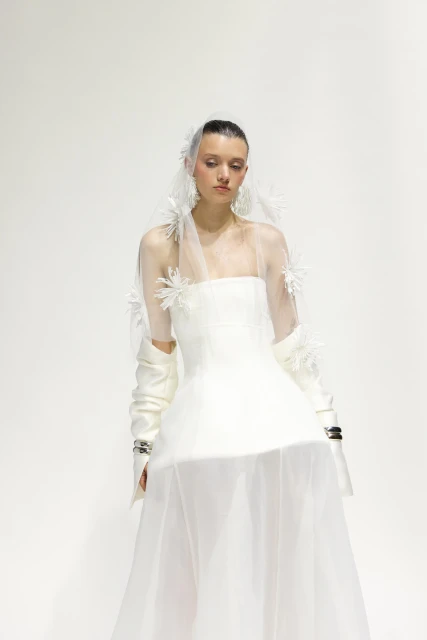 A woman poses in an off-shoulder white wedding dress and tulle veil with white flower accents against a plain white backdrop.
