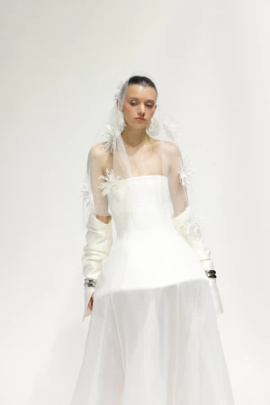 A woman poses in an off-shoulder white wedding dress and tulle veil with white flower accents against a plain white backdrop.