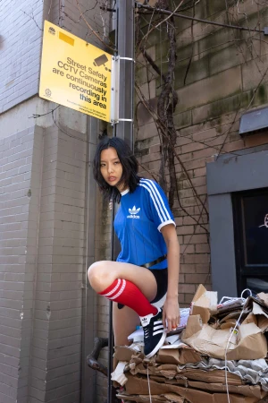 A young woman poses on a pile of cardboard boxes in an urban setting, wearing a blue Adidas shirt, black shorts, and red and white socks.