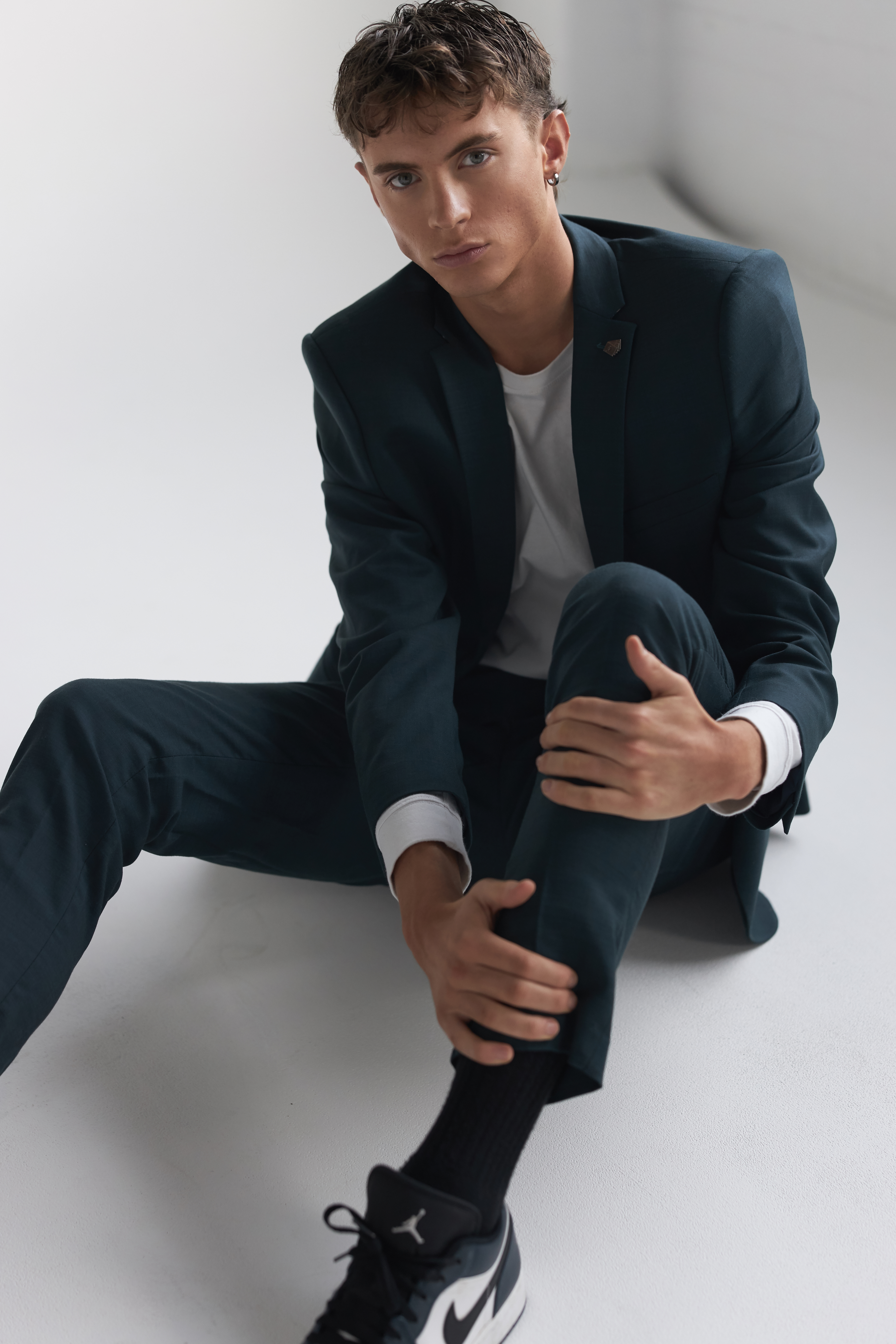 Young adult male in dark suit and sneakers sitting on a white surface looking at the camera, wearing a white t-shirt and dark socks.