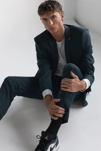 Young adult male in dark suit and sneakers sitting on a white surface looking at the camera, wearing a white t-shirt and dark socks.