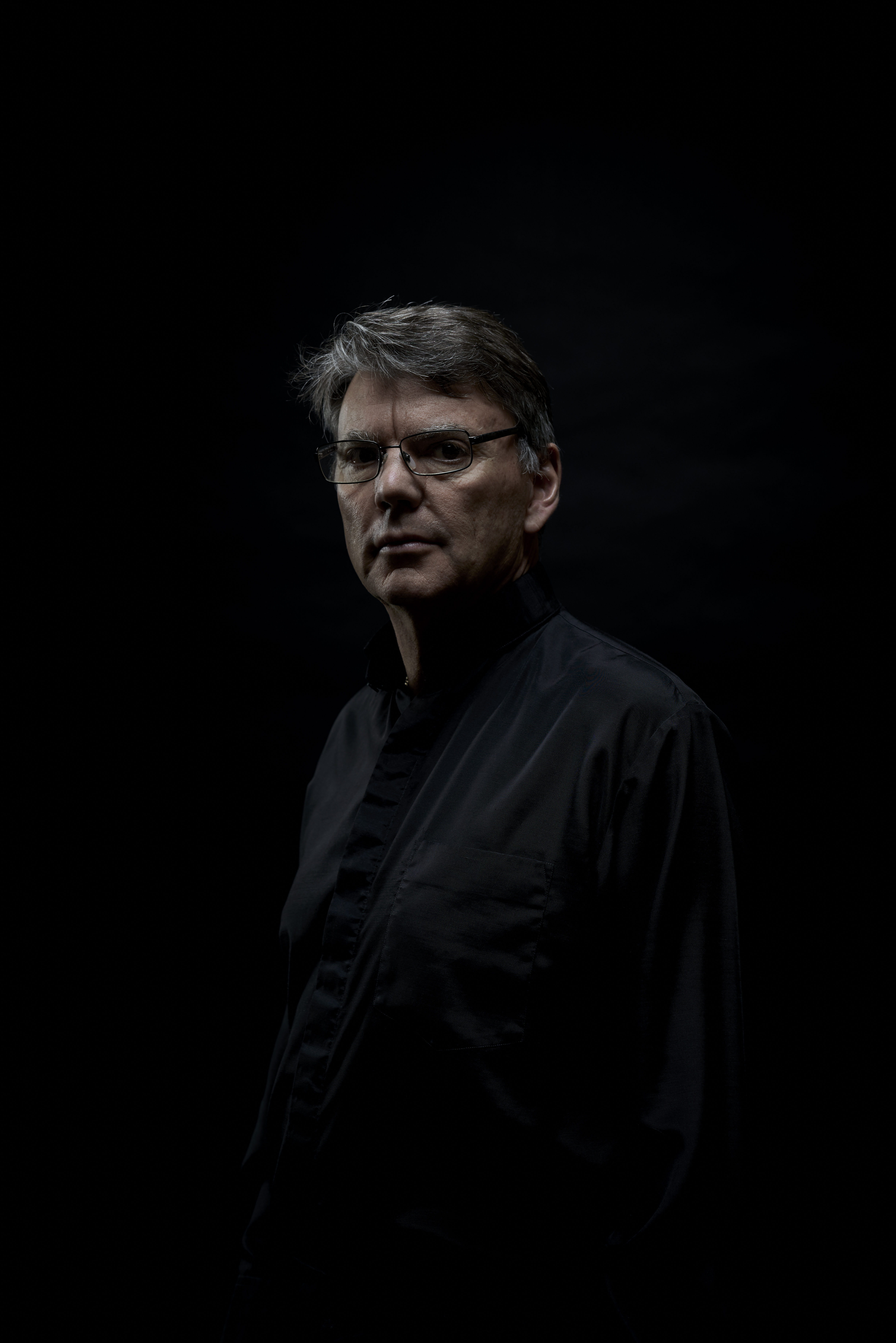 Portrait of a mature man wearing glasses and a dark shirt, looking directly at the camera against a dark background with dramatic lighting.