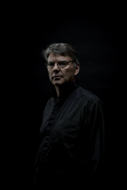 Portrait of a mature man wearing glasses and a dark shirt, looking directly at the camera against a dark background with dramatic lighting.