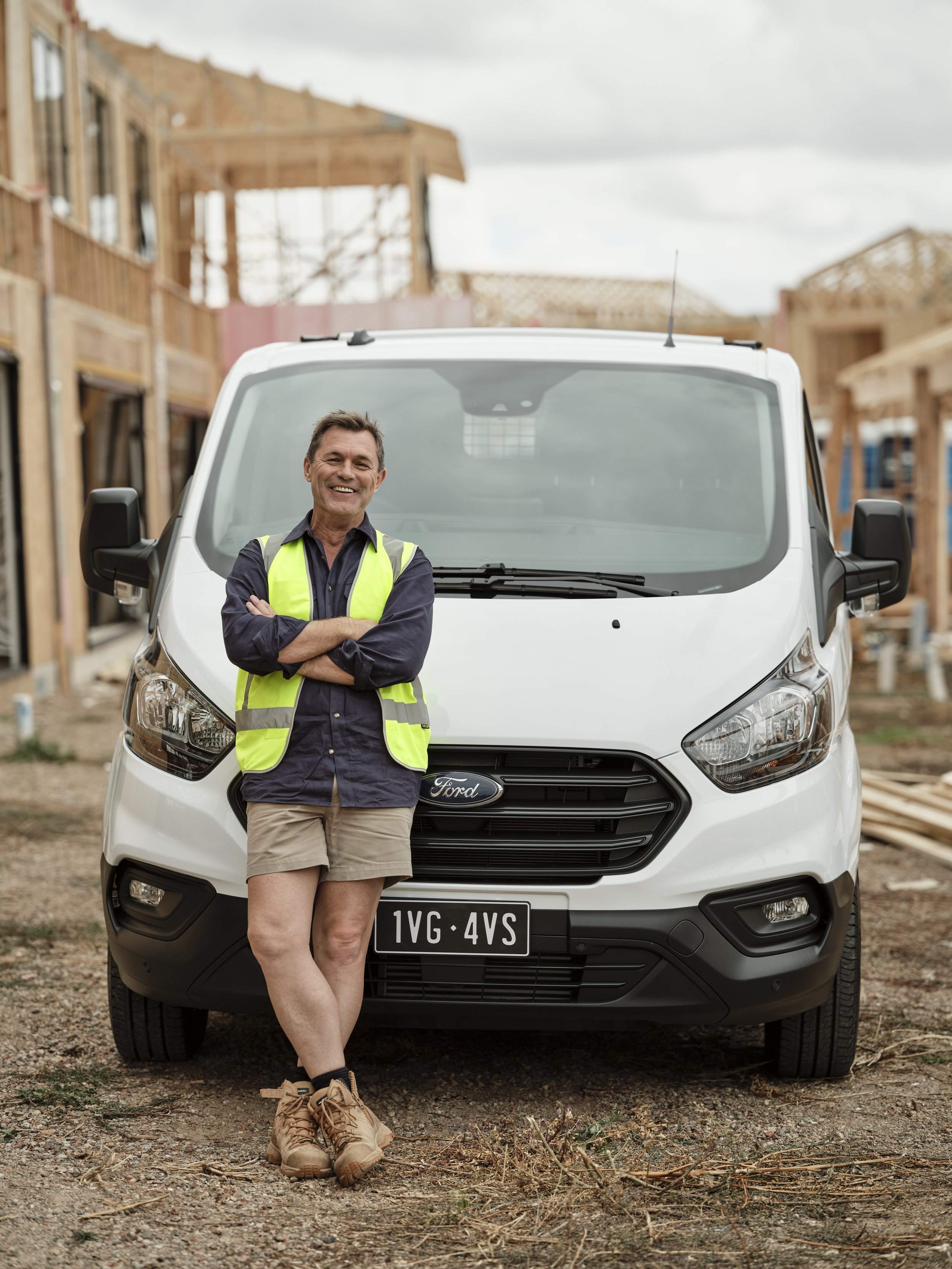 Man wearing a safety vest and work boots poses in front of a white van at a construction site on a cloudy day. 