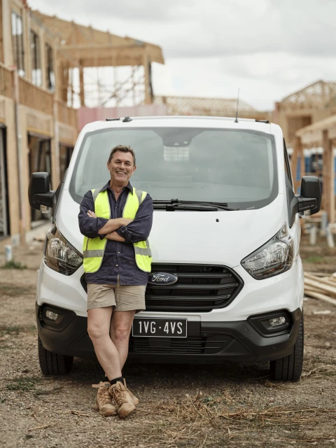 Man wearing a safety vest and work boots poses in front of a white van at a construction site on a cloudy day. 