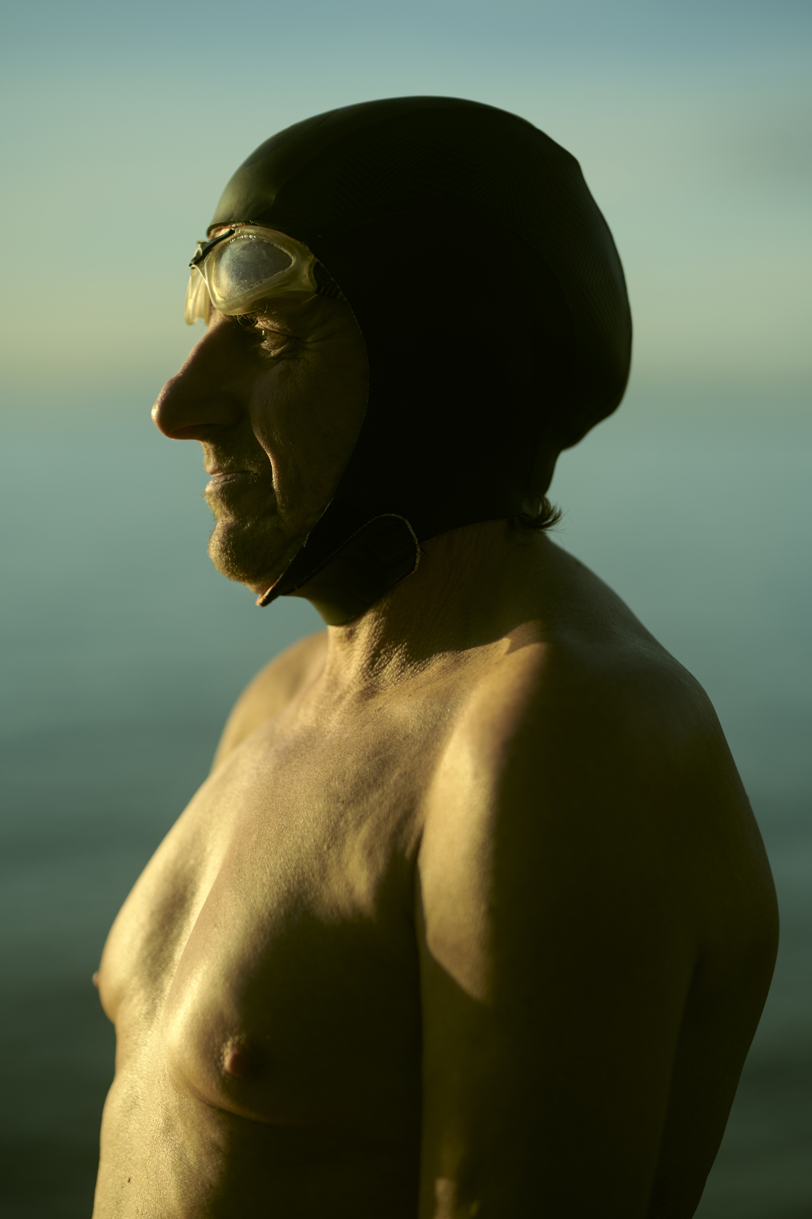 Profile of older man wearing swim cap and goggles, shirtless, in front of water. Golden light illuminating the swimmer. 