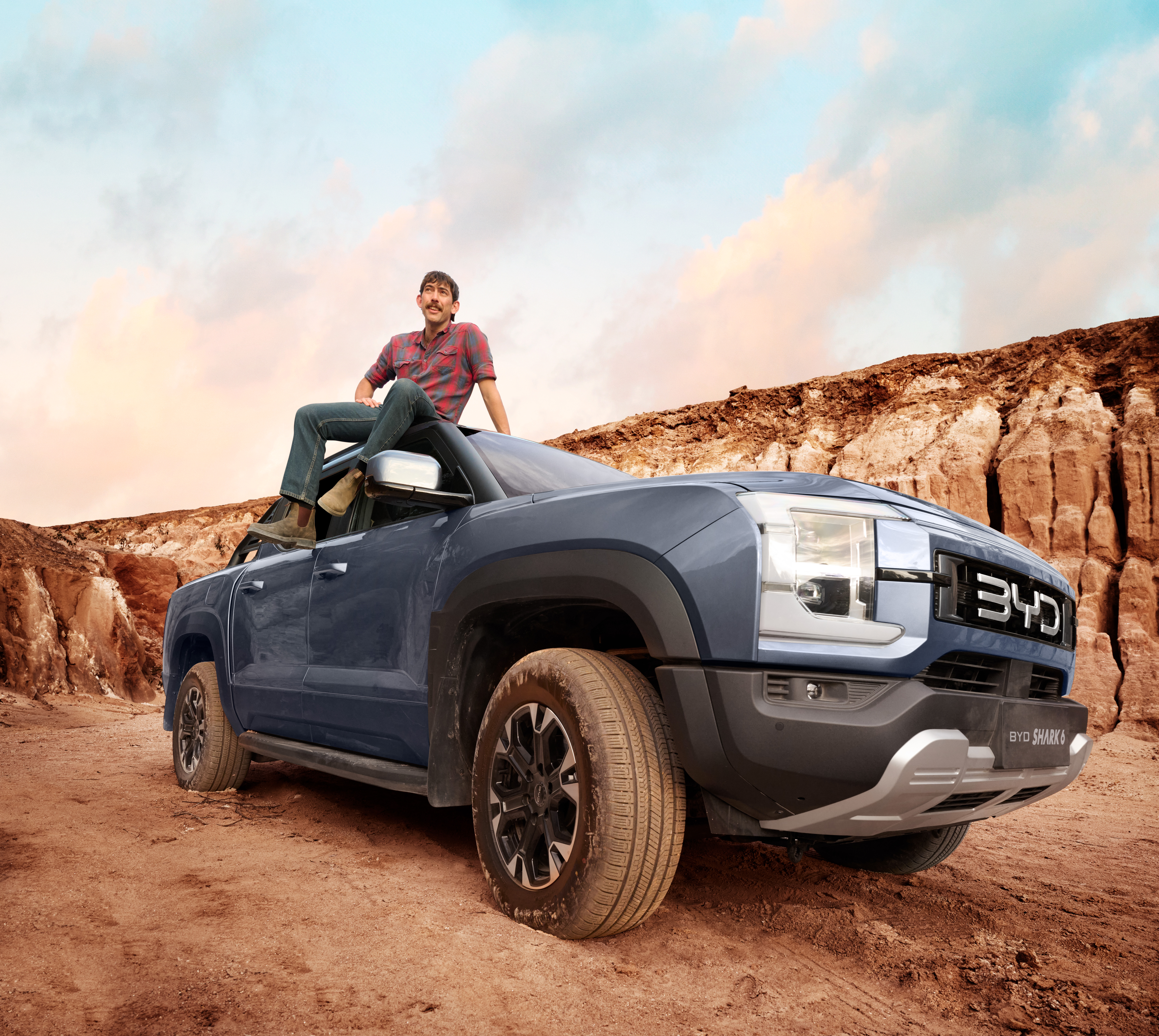 Man poses on a blue BYD Shark pickup truck in a desert landscape under a blue and cloudy sky; automotive product shot.