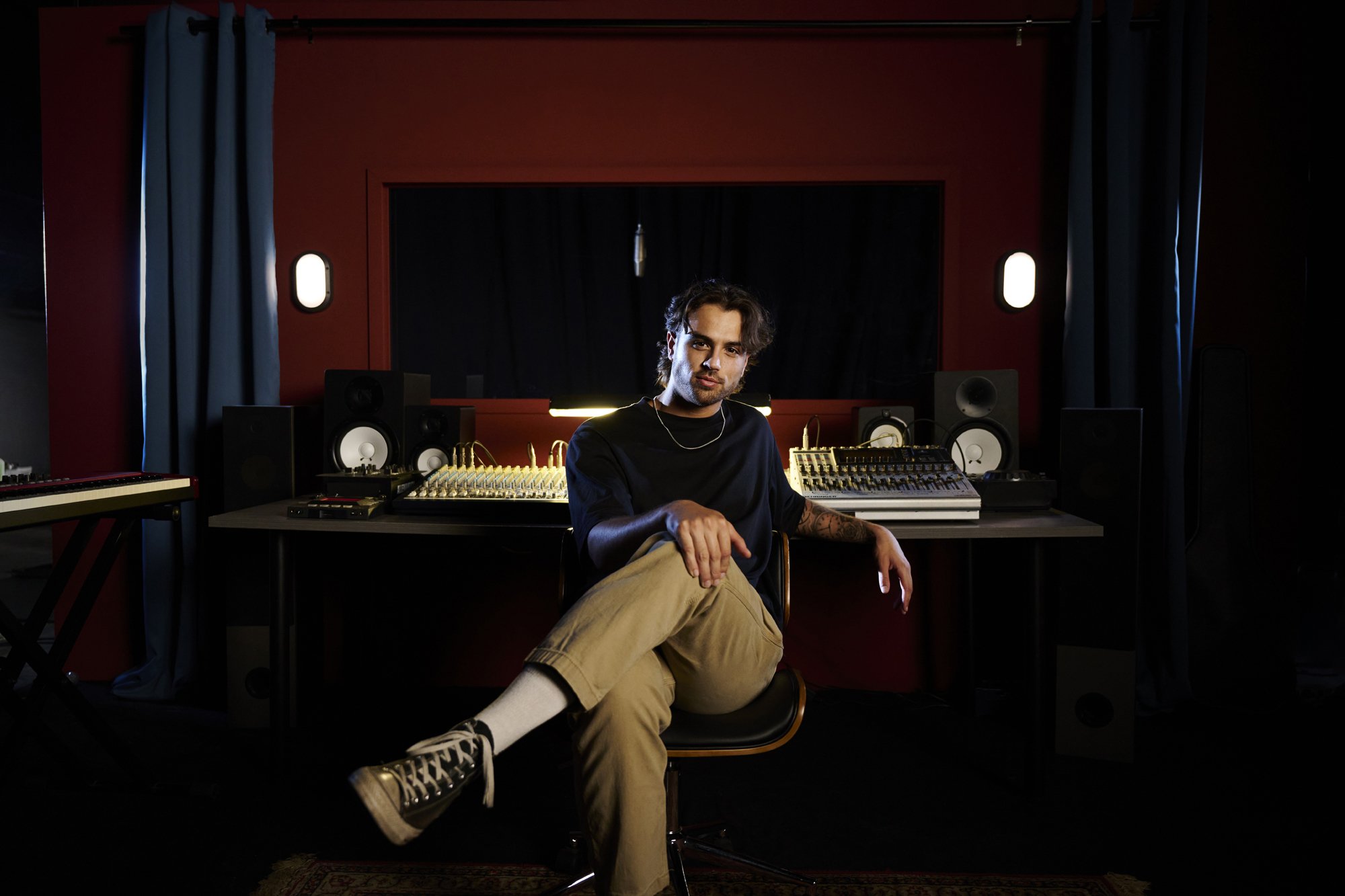 Adult male sits in a music studio with a soundboard, speakers, microphone, and instrument keyboard. Dark red walls and curtains.