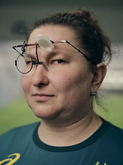 Close-up portrait of a woman wearing shooting glasses, indoors. She wears a green shirt with a yellow logo.