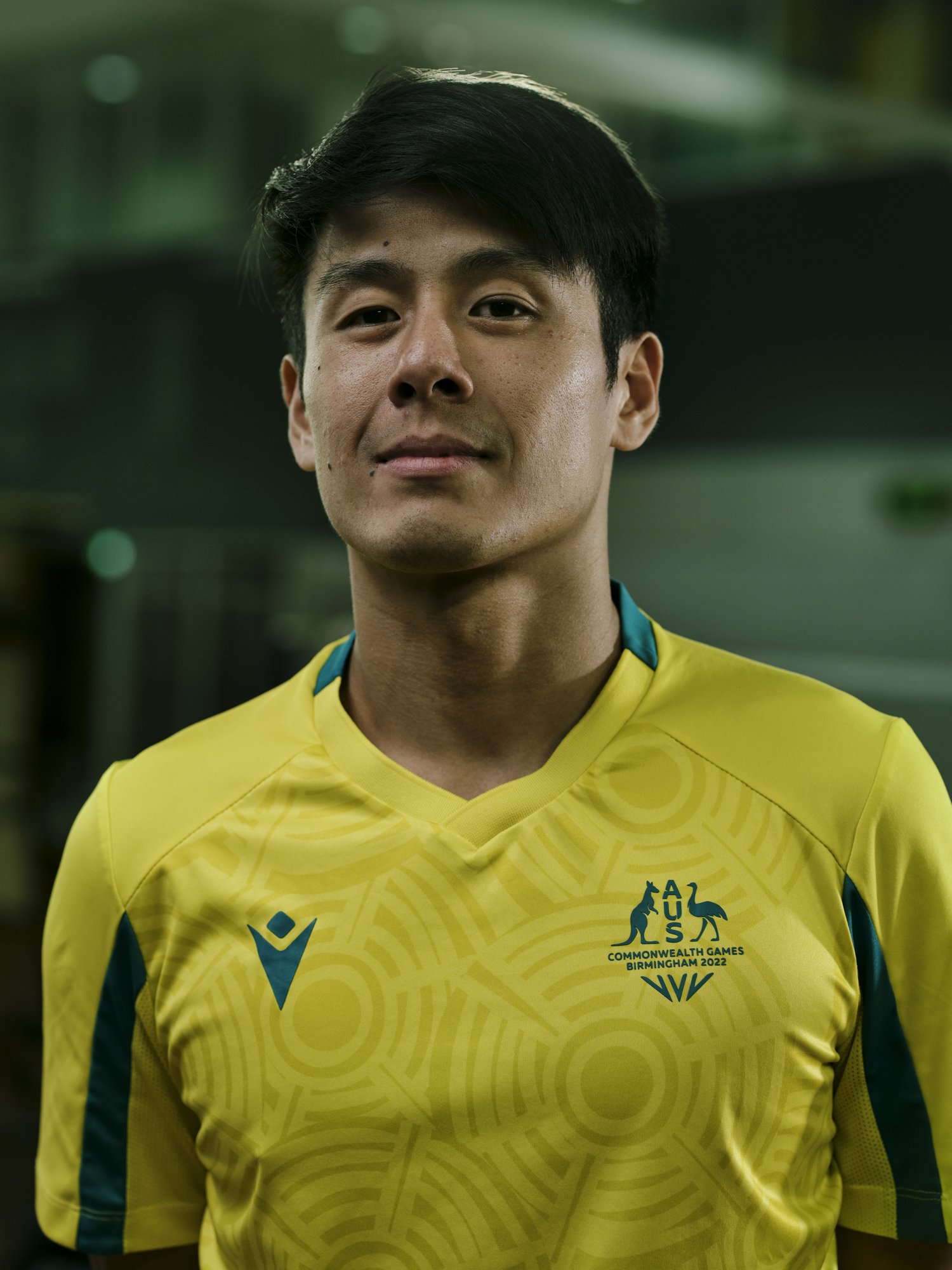 Portrait of Asian male athlete in yellow and green uniform with Commonwealth Games logo, smiling slightly against blurred backdrop.