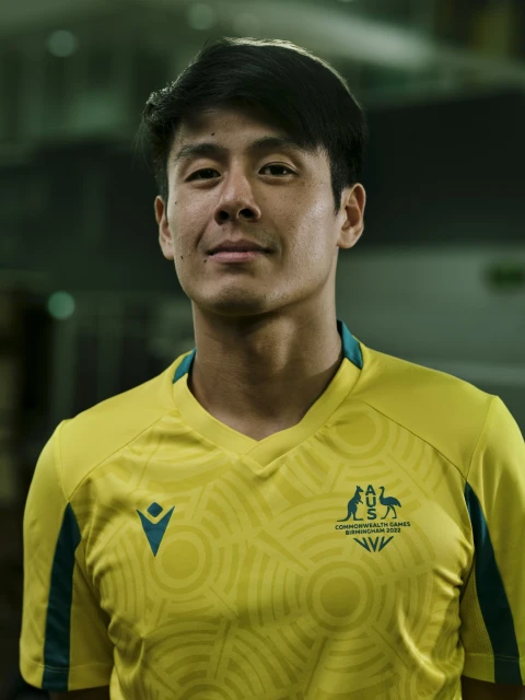 Portrait of Asian male athlete in yellow and green uniform with Commonwealth Games logo, smiling slightly against blurred backdrop.