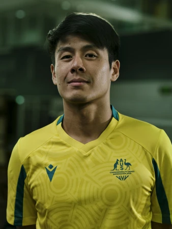 Portrait of Asian male athlete in yellow and green uniform with Commonwealth Games logo, smiling slightly against blurred backdrop.