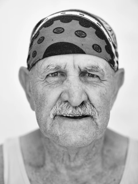 Monochrome portrait of an elderly man with a mustache wearing a patterned bandana, neutral facial expression, in front of a white background.