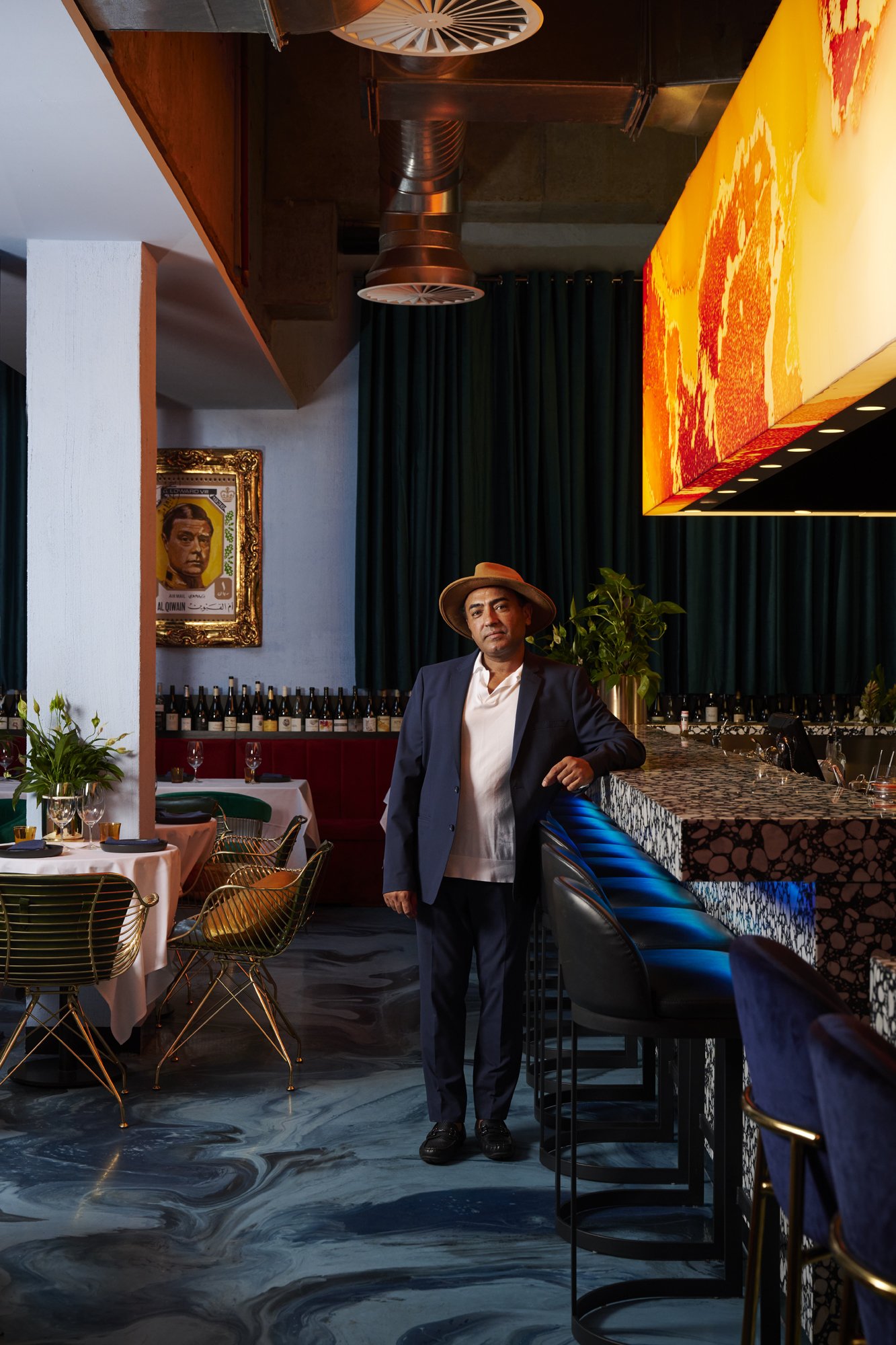 Man in a suit and hat stands inside a restaurant with a blue and brown marbled floor, tables with wine, and abstract overhead lighting.