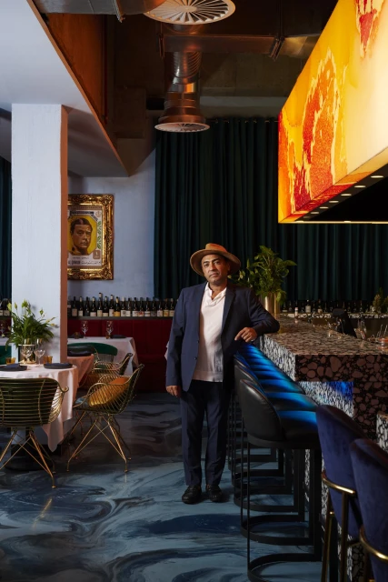 Man in a suit and hat stands inside a restaurant with a blue and brown marbled floor, tables with wine, and abstract overhead lighting.