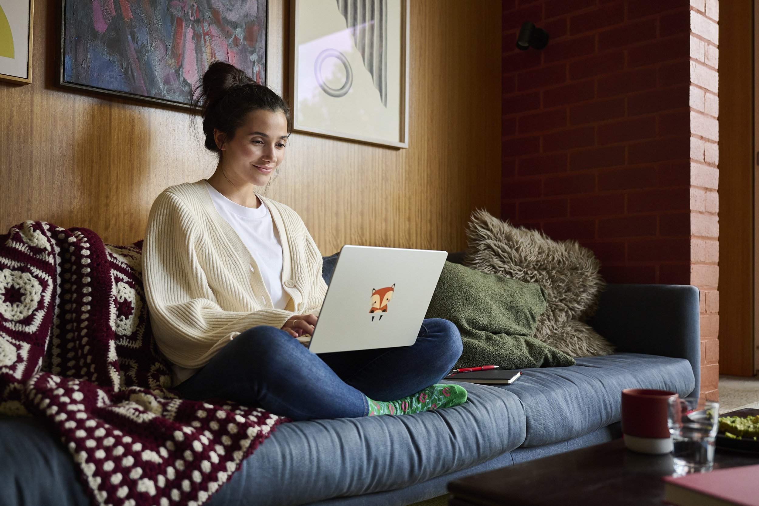Woman sitting cross-legged on a couch using a laptop with a fox sticker, photographed by Michael Oulton.