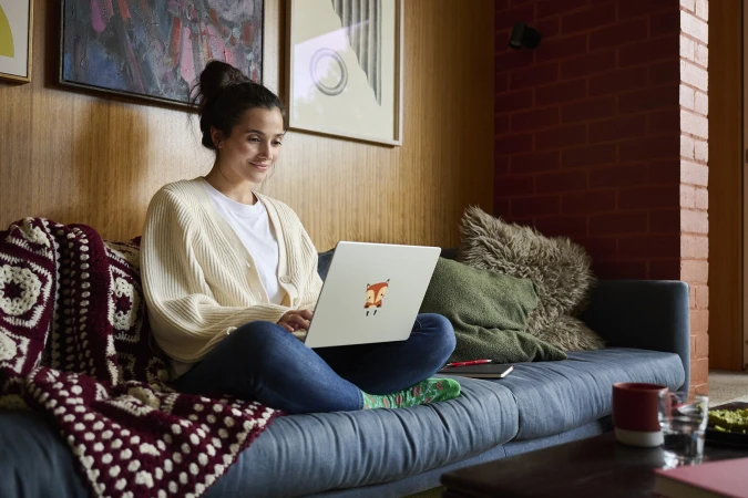 Woman sitting cross-legged on a couch using a laptop with a fox sticker, photographed by Michael Oulton.