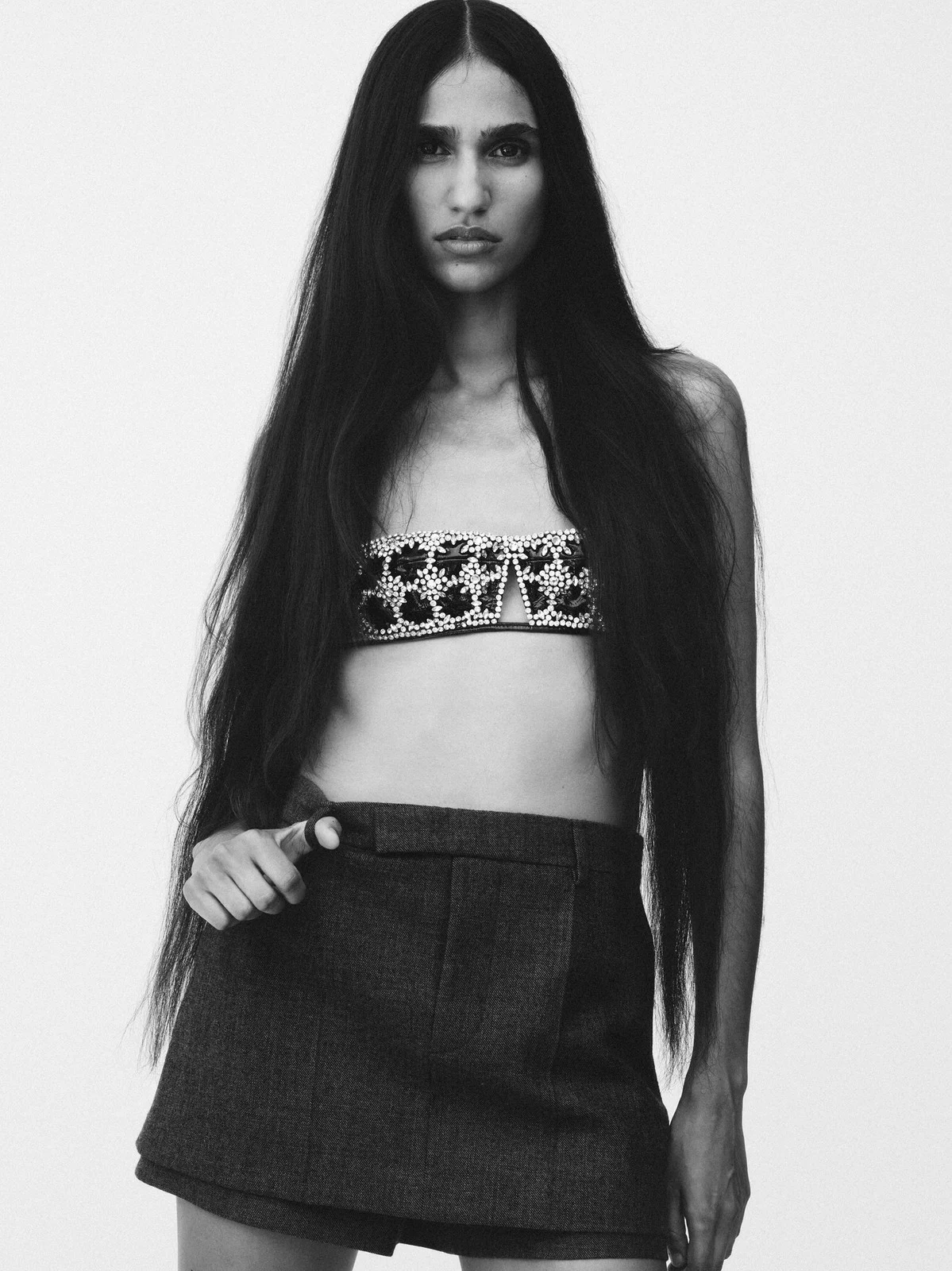 Black and white portrait of a woman with long dark hair wearing a beaded top and skirt against a light background in a studio setting.