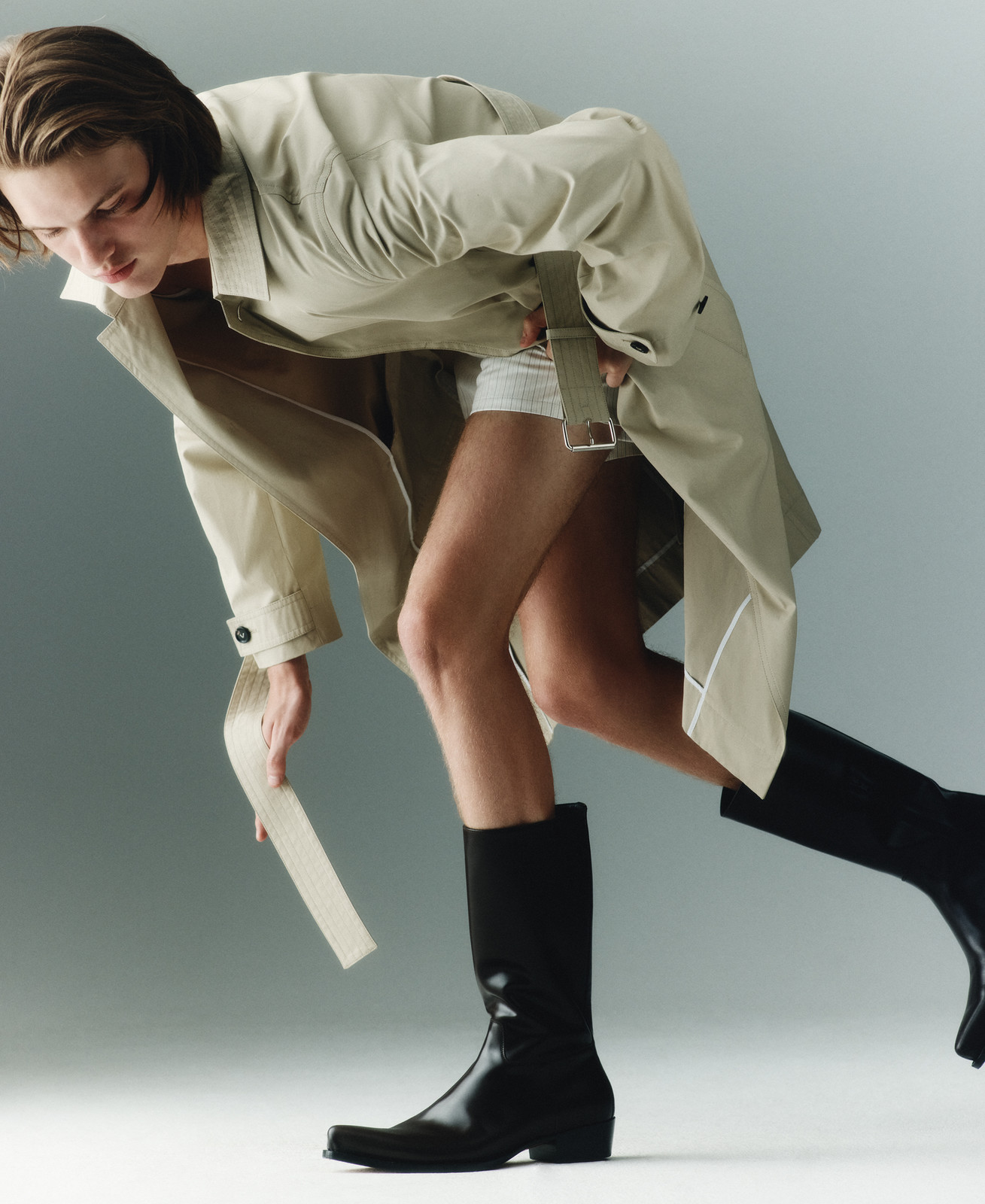 Male fashion model strikes a pose in a studio setting wearing a beige trench coat, white shorts, and black riding boots against a grey background.