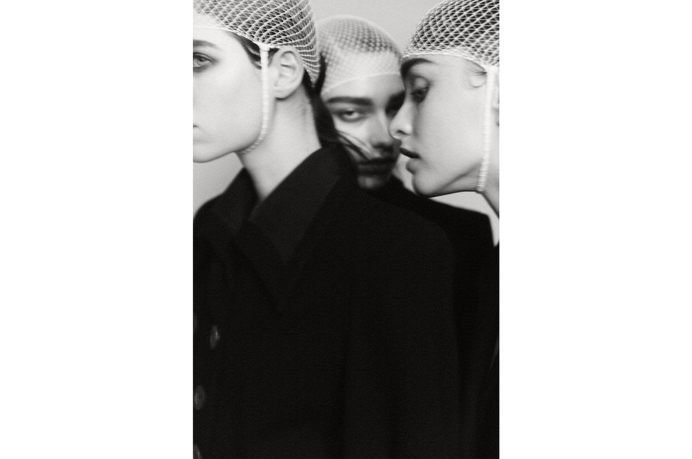 Monochrome portrait of three women with netted caps, dark coats against white background, evokes high-fashion editorial aesthetic.