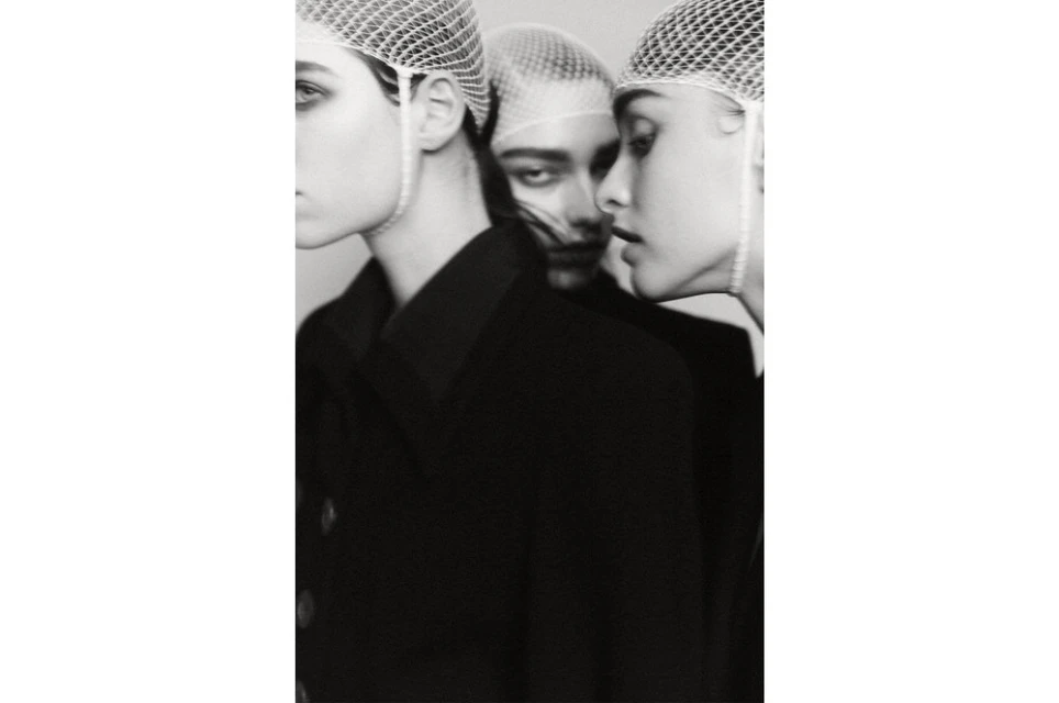 Monochrome portrait of three women with netted caps, dark coats against white background, evokes high-fashion editorial aesthetic.